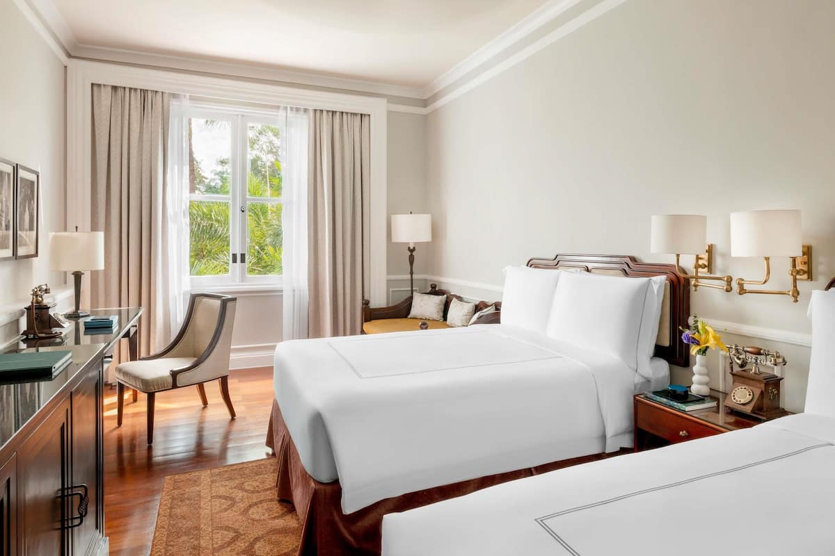 A hotel room with two beds, a desk with a lamp, a chair, a nightstand with a lamp, and a window with curtains.
