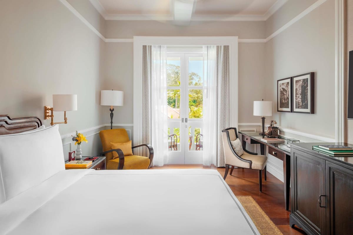 A hotel room with a large bed, a desk, two chairs, a wardrobe, and a view through a glass door.