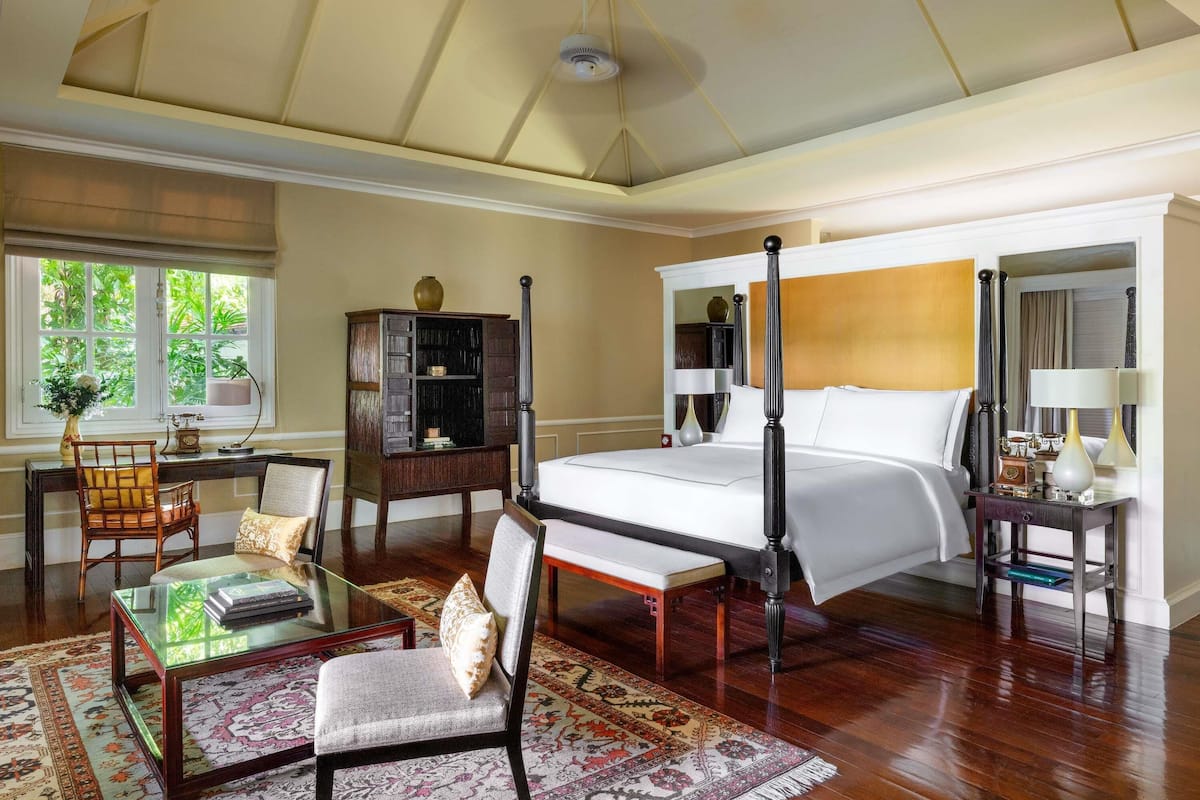 A bedroom with a four-poster bed, a desk with a chair, a bookshelf, and a large window with a view of greenery.