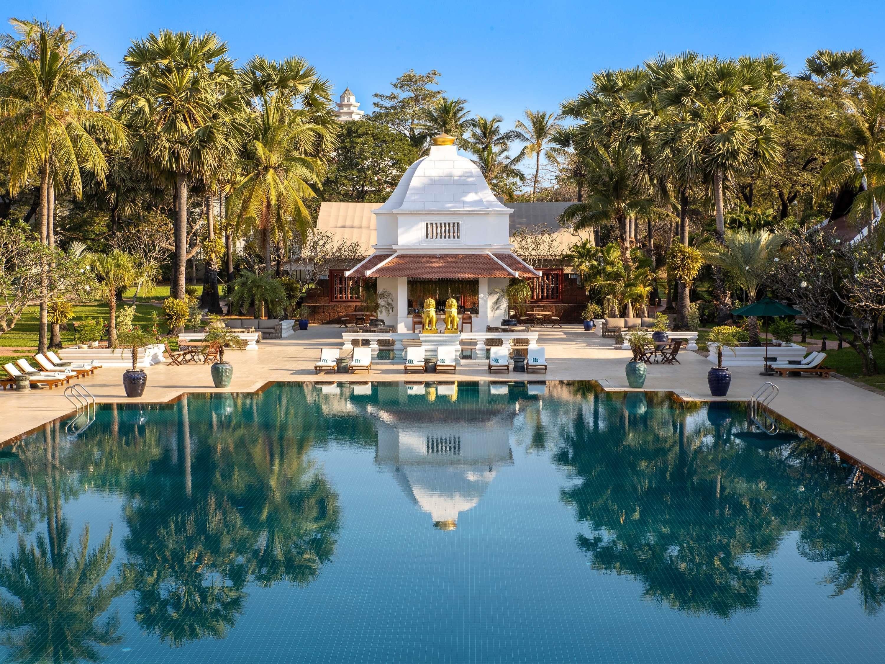 Uma piscina com espreguiçadeiras, um edifício branco com cúpula e palmeiras.
