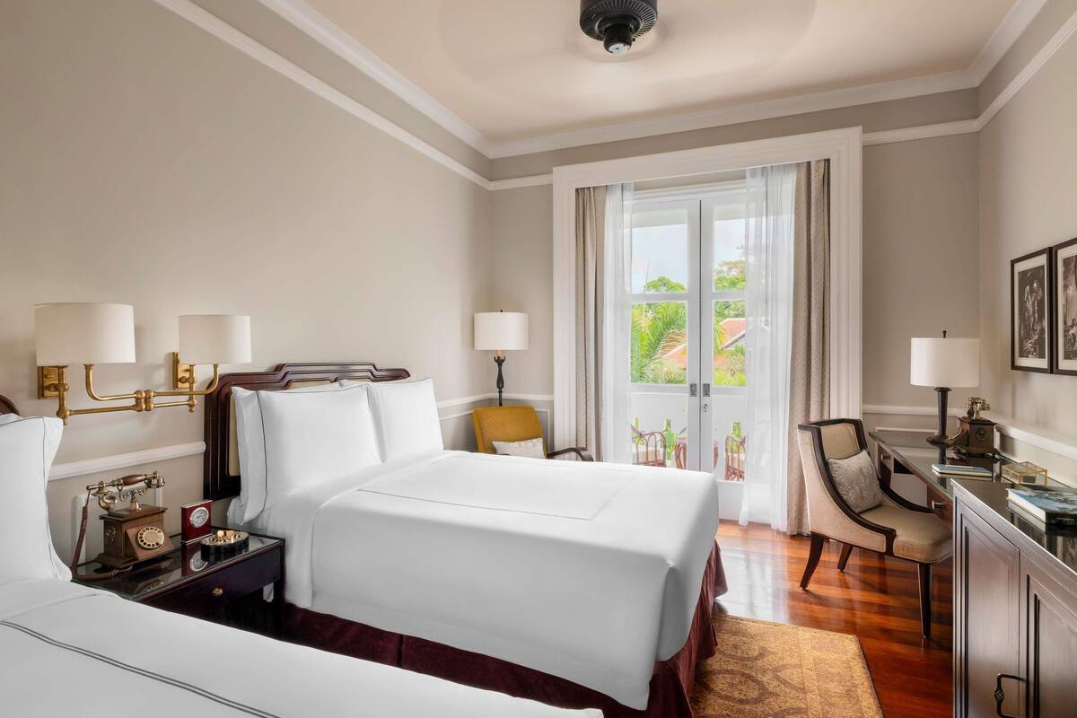 A hotel room with a large bed, a desk with a chair, a desk lamp, and a view of the outdoors through a glass door.