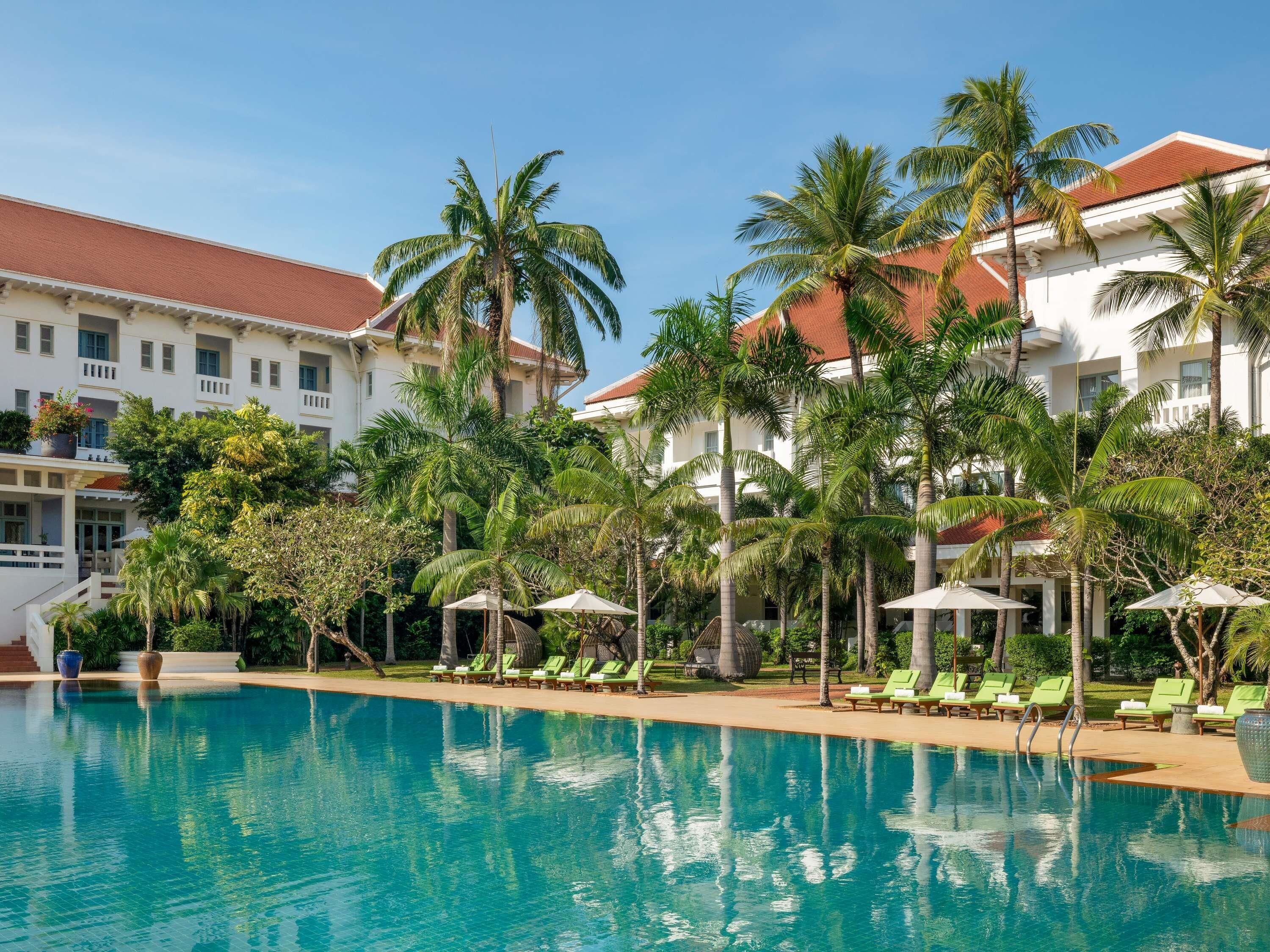 A pool area with lounge chairs and umbrellas, surrounded by palm trees and a building with a red roof.