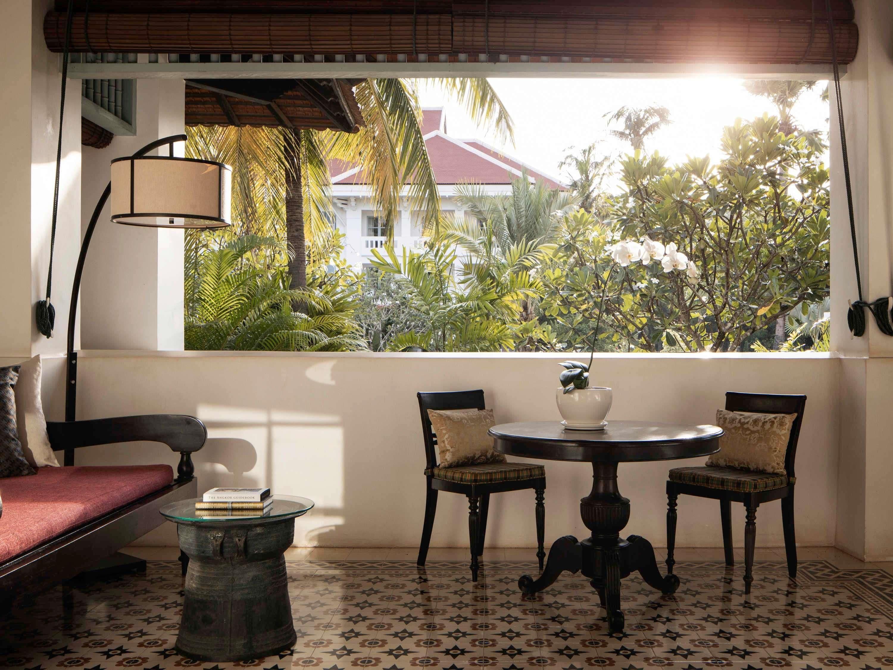A cozy room with a view of lush greenery and a red-roofed building. There is a round table with two chairs, a small side table, and a sofa with cushions.