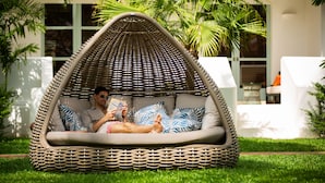 A wicker lounge chair with a cushion, a person reading a book, and a lush green lawn.