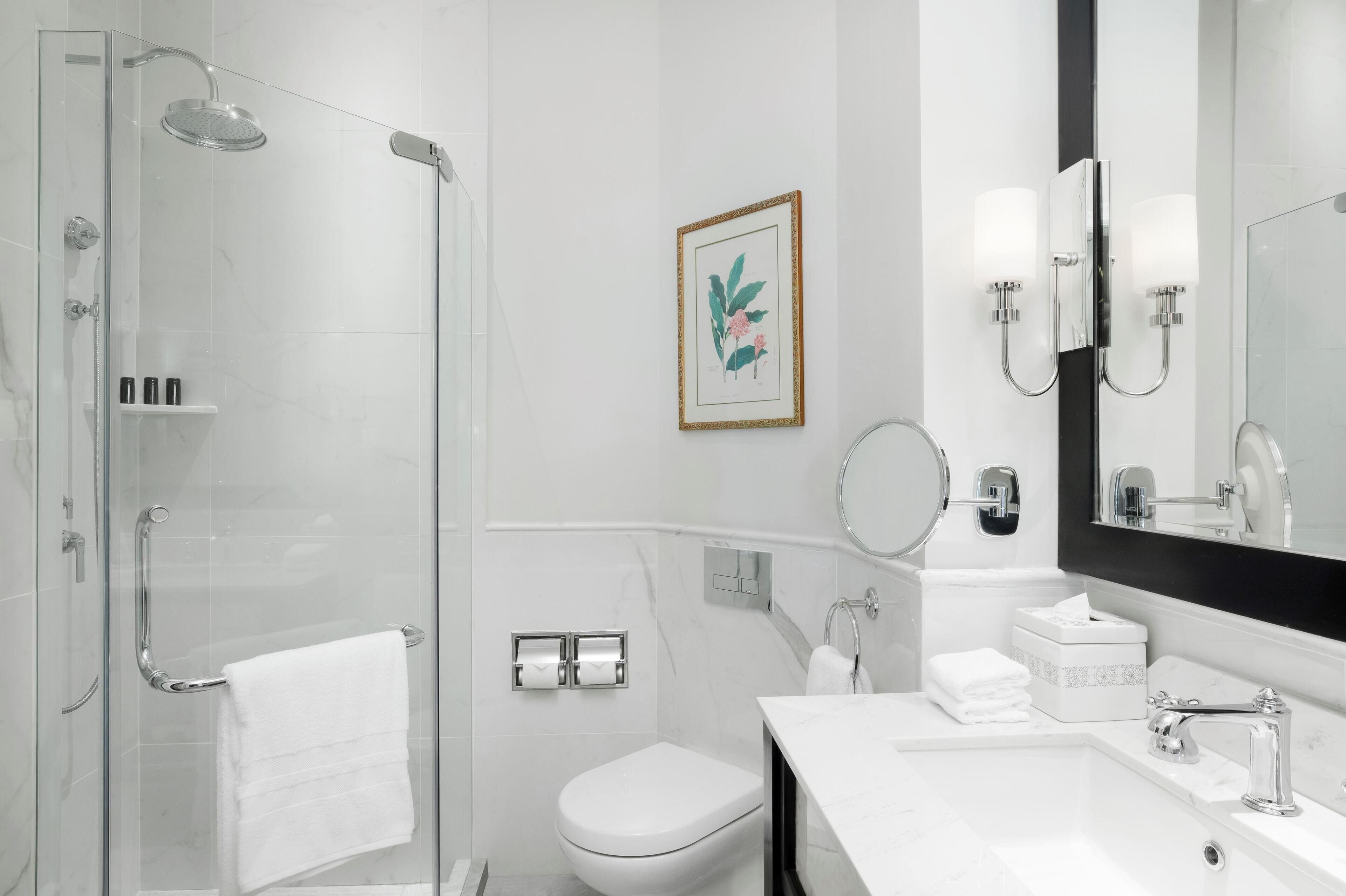 Een moderne badkamer met een douchecabine van glas, een wit toilet, een wastafel met spiegel en een lijst met een botanische print aan de muur.