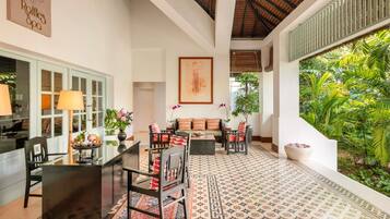 A spacious room with a high ceiling, a dining area with a table and chairs, a sofa, and a large window overlooking a lush garden.