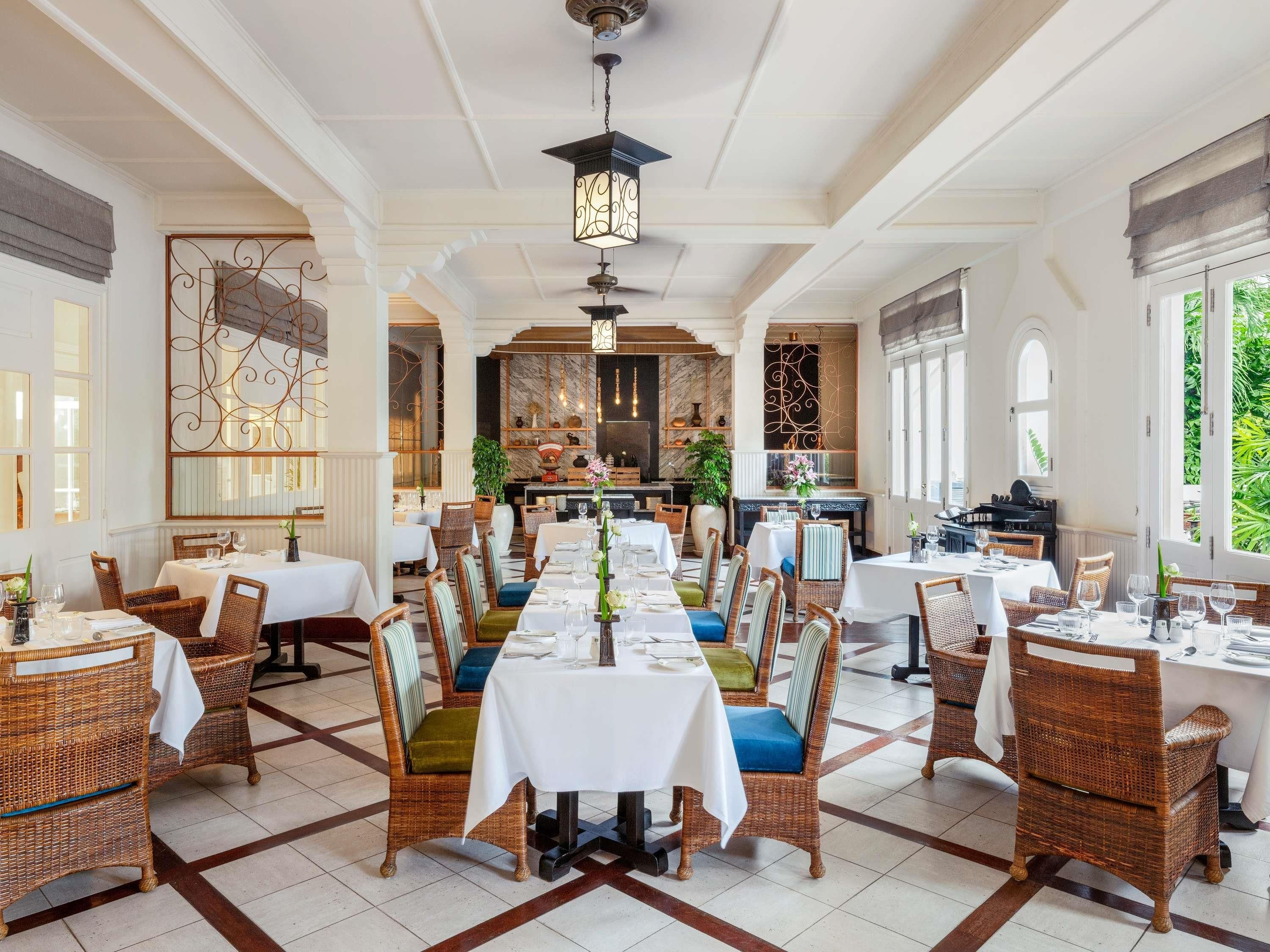 A spacious dining area with multiple tables and wicker chairs, a kitchen in the background, and large windows.