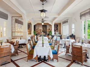A spacious dining area with multiple tables and wicker chairs, a kitchen in the background, and large windows. - Raffles Grand Hotel d'Angkor (Siem Reap)