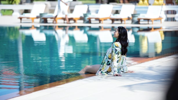 A woman sitting by a pool with lounge chairs and golden statues in the background.
