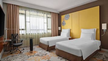 Ein Hotelzimmer mit zwei Betten, einem Schreibtisch mit Stuhl, einem Fernseher und einem großen Fenster mit Vorhängen.