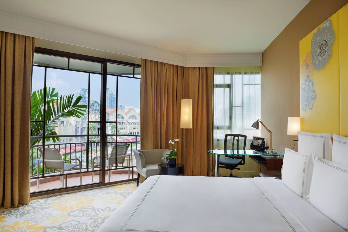 A hotel room with a large bed, a desk with a chair, a small table with a lamp, and a view of the city through large windows.