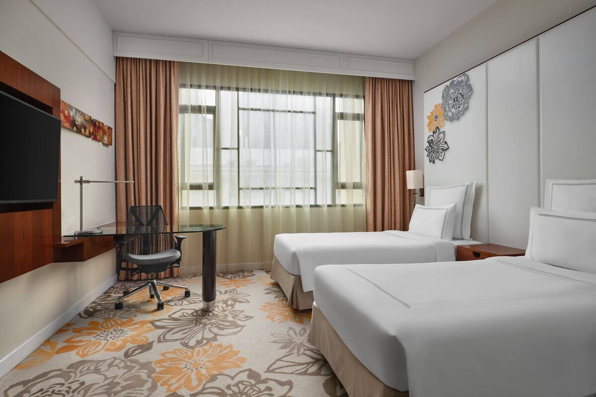 A hotel room with two beds, a desk with a chair, a TV, and a window with curtains.