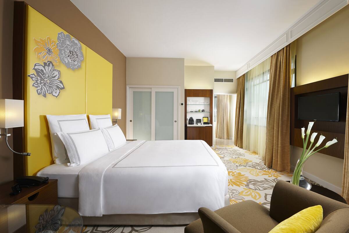 A hotel room with a large bed, a desk with a lamp, a TV, and a decorative wall piece.
