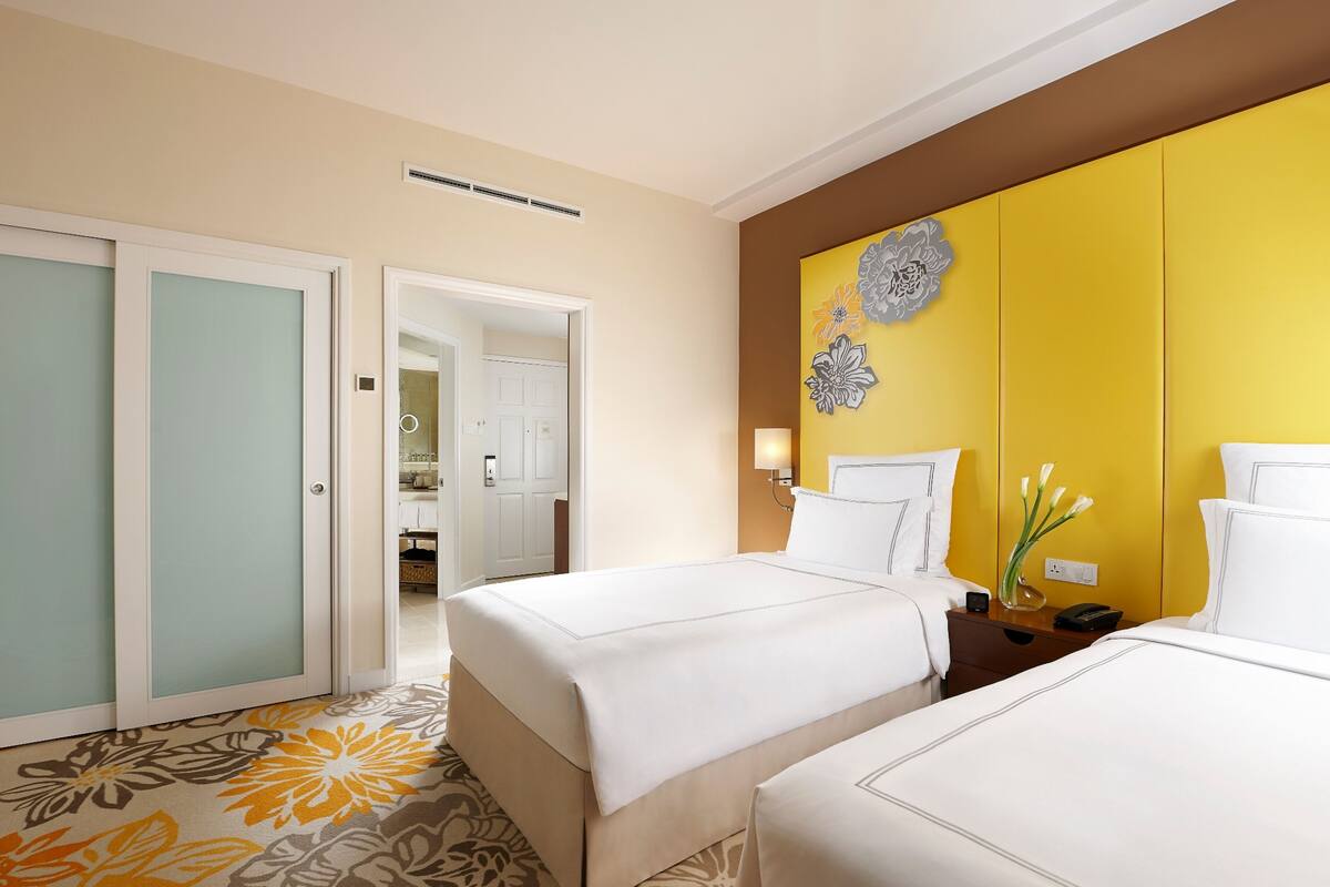 A hotel room with two beds, a bathroom visible through an open door, and a yellow accent wall with a floral design.