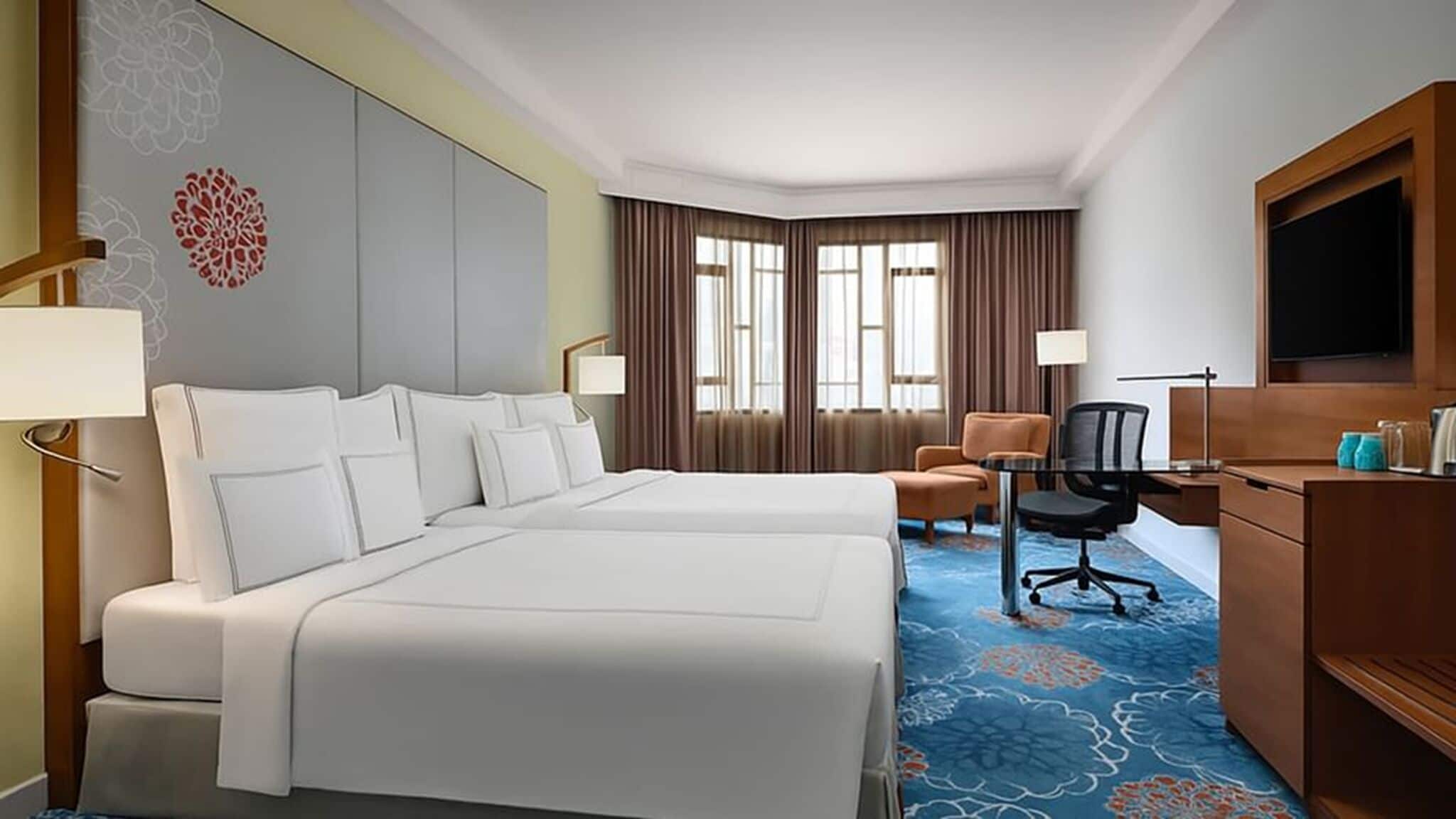 A hotel room with two beds, a desk, a chair, a TV, and a large window with curtains.