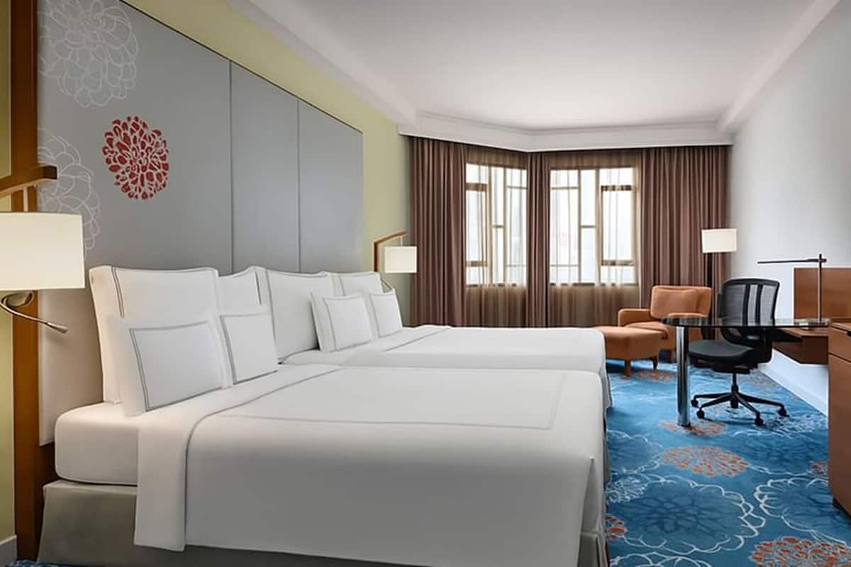 A hotel room with two beds, a desk, a chair, a TV, and a large window with curtains.