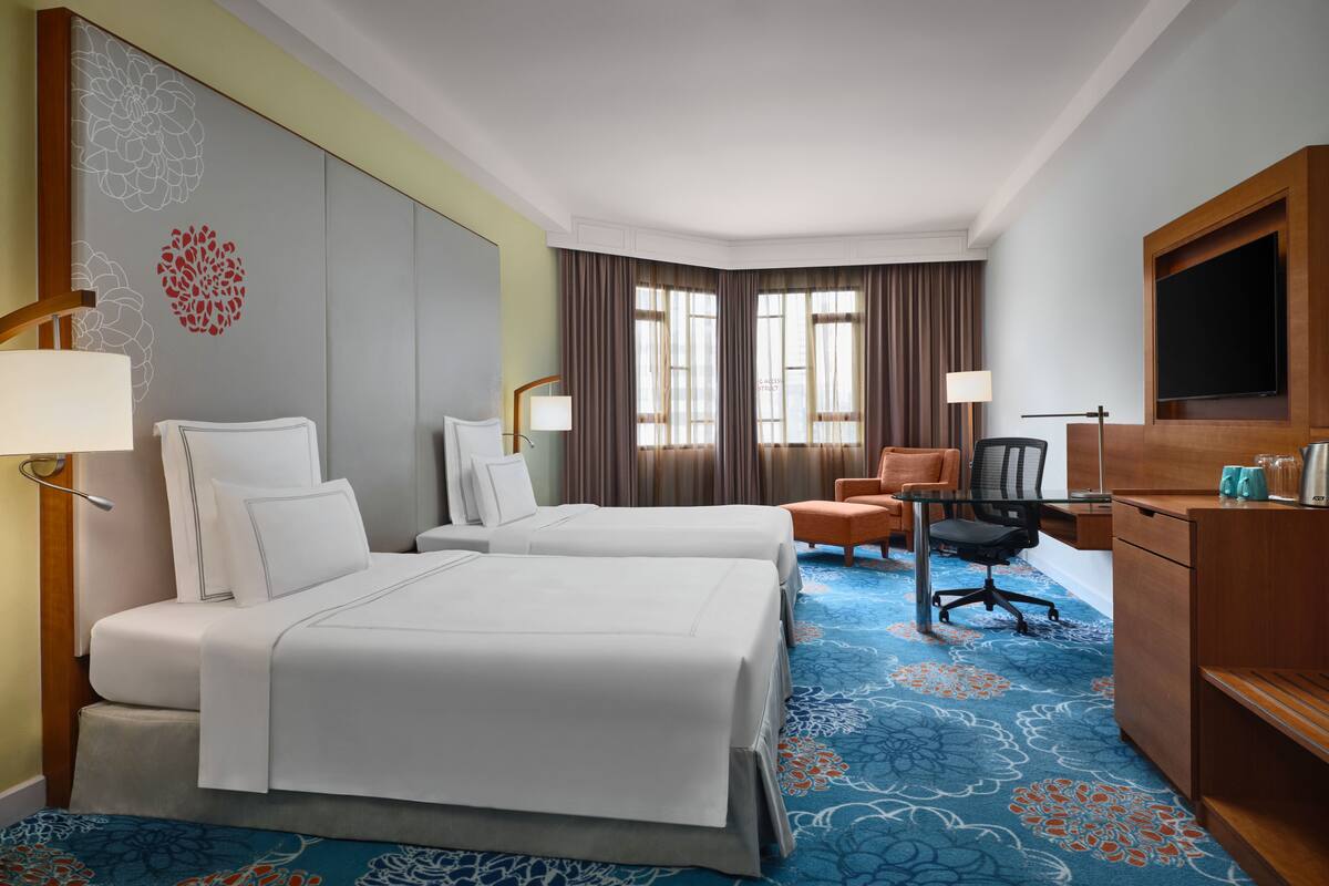 A hotel room with two beds, a desk, a chair, a TV, and a window with curtains.