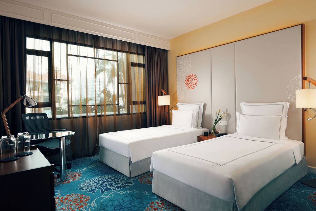 A hotel room with two beds, a desk, a chair, a lamp, and a view of palm trees through the window.
