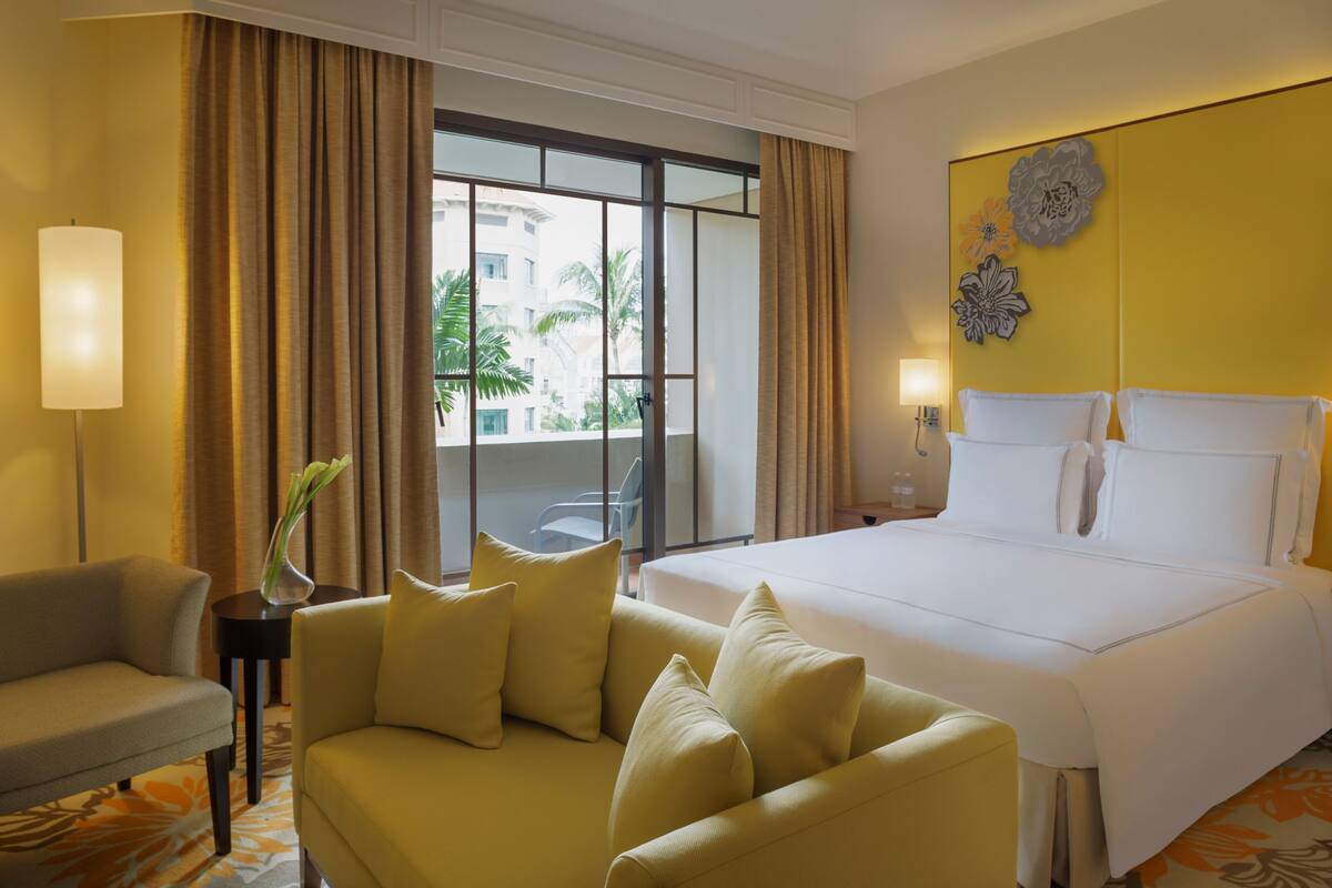 A hotel room with a large bed, a sofa, a small table with a vase, and a view of palm trees outside.