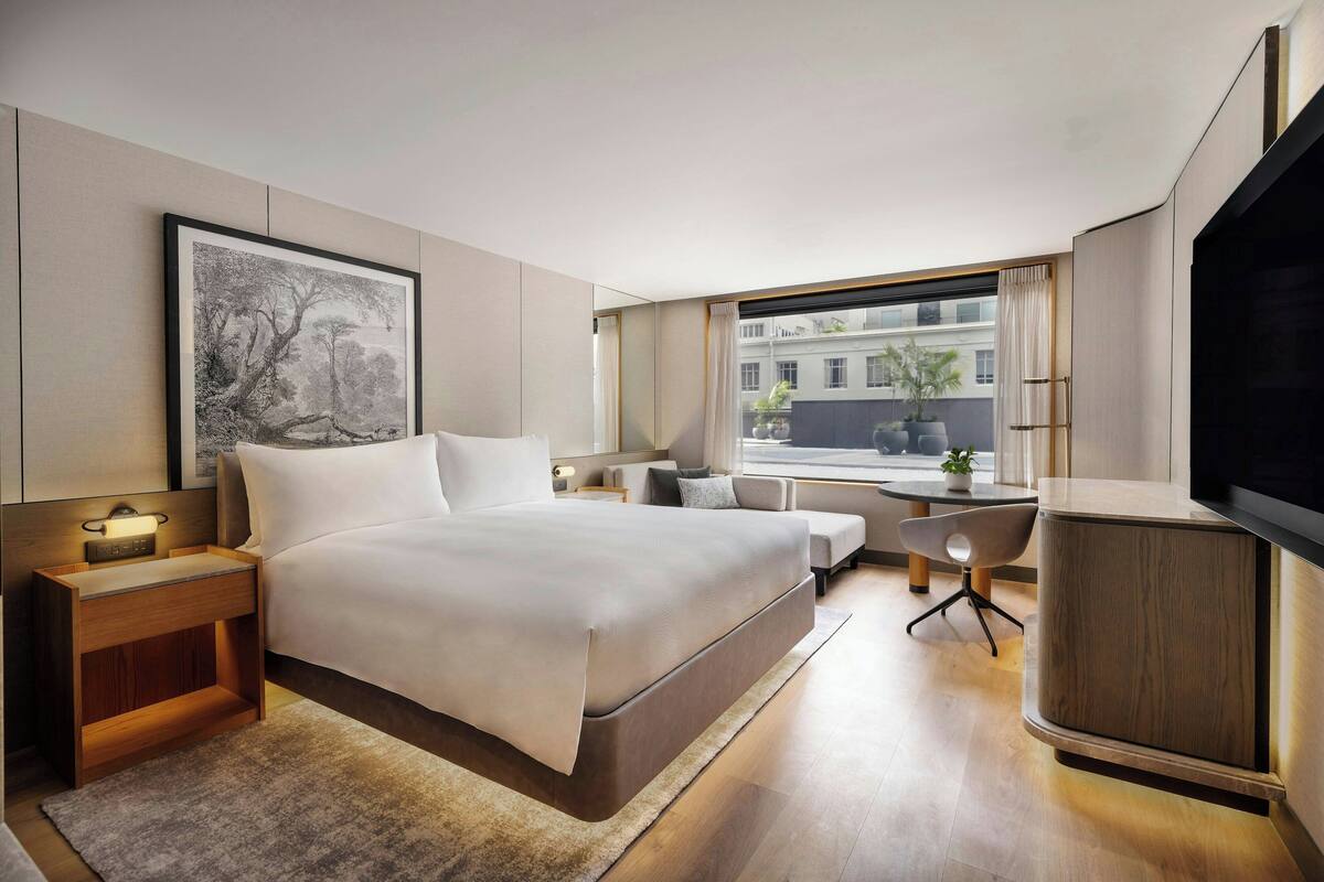 A modern hotel room with a large bed, bedside table with a lamp, a desk, a chair, and a TV.
