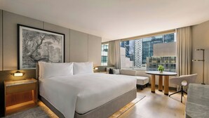 Une chambre d’hôtel moderne dotée d’un grand lit, de tables de chevet, d’un petit bureau et offrant une vue sur la ville grâce à de grandes fenêtres.