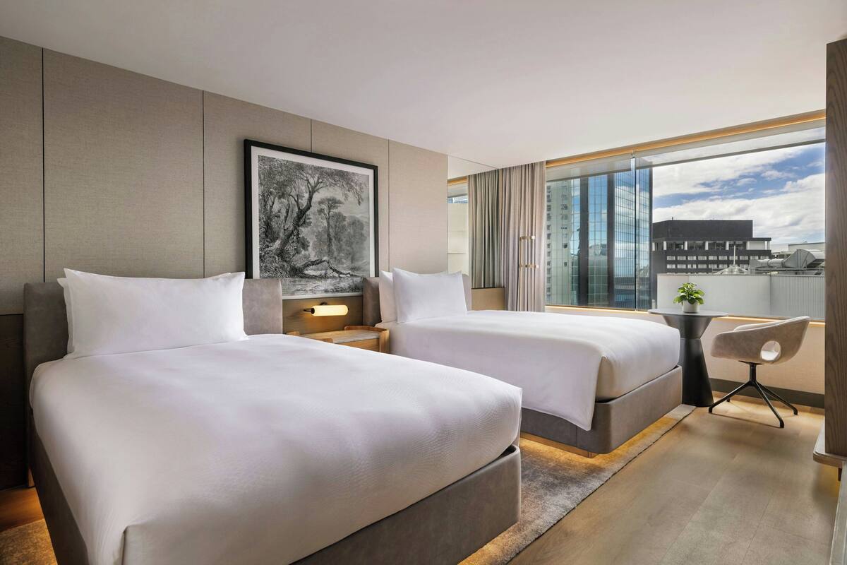 A hotel room with two beds, a small table, a chair, a large window with a city view, and a framed picture on the wall.