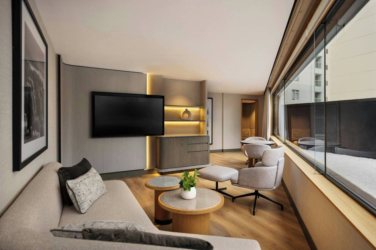 A modern living room with a sofa, round coffee table, and armchairs. A flat-screen TV is mounted on the wall, and there is a window with a view of the city.