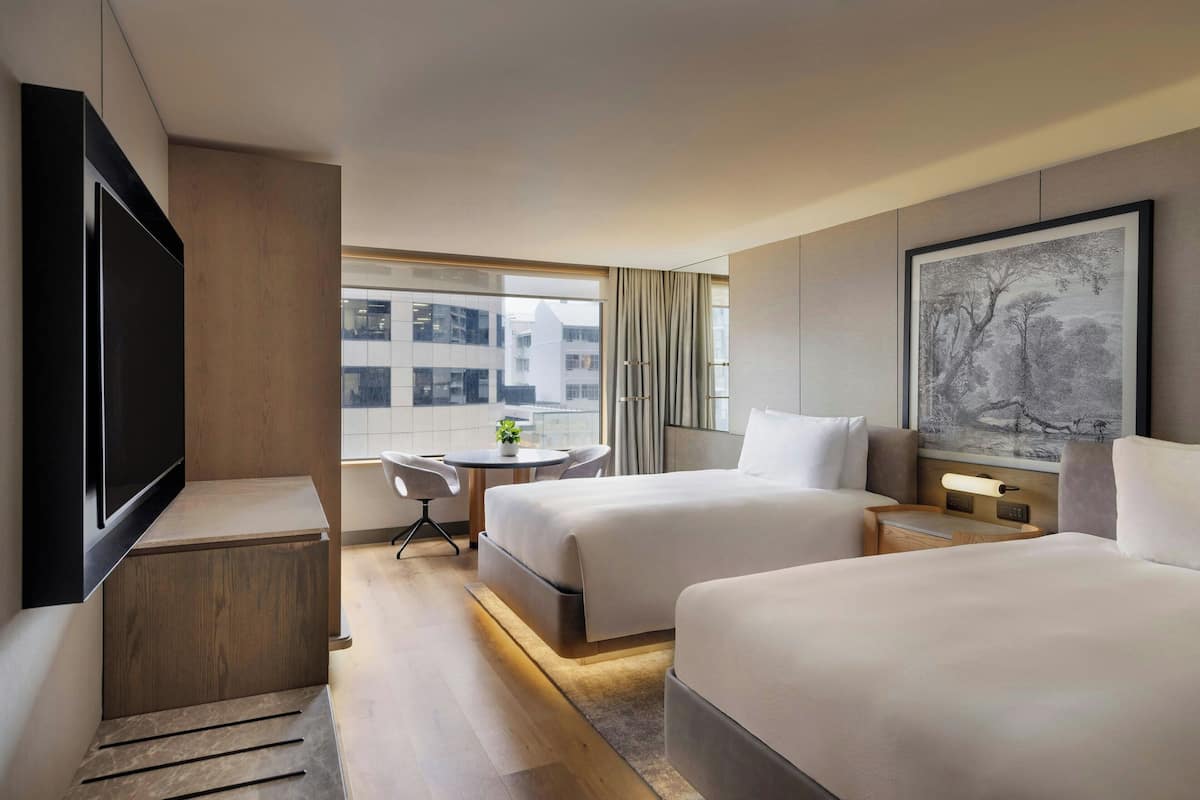 A hotel room with two beds, a TV, a desk, a chair, and a window with a view of buildings.