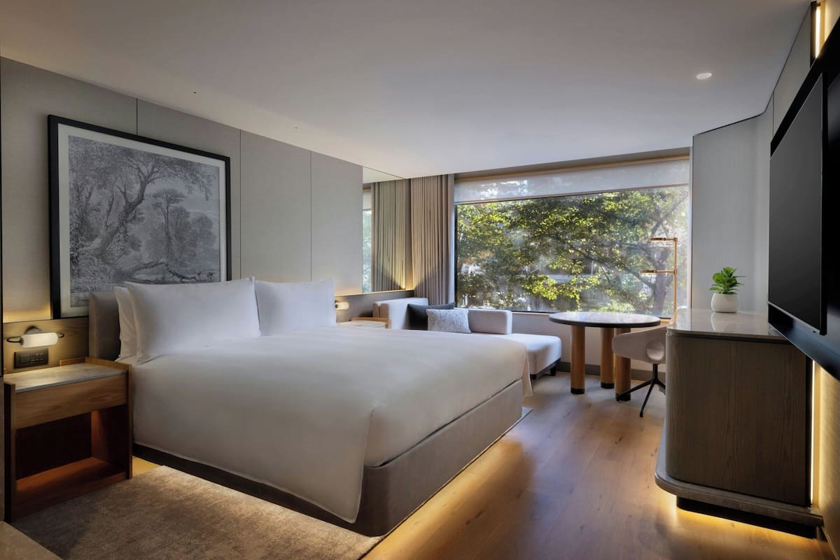 A modern hotel room with a large bed, a desk, a chair, a small table, and a TV.