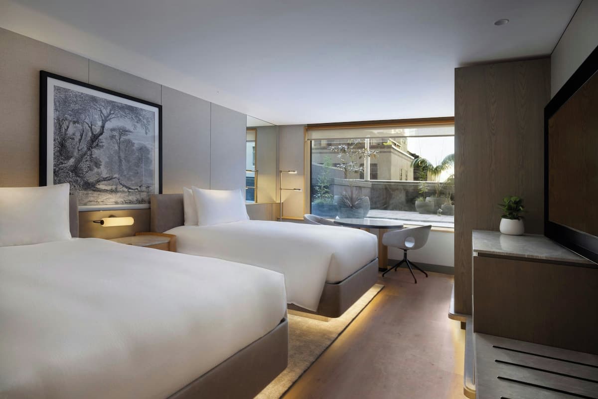 A modern hotel room with two beds, a desk, a chair, a TV, and a large window offering a view of the outside.