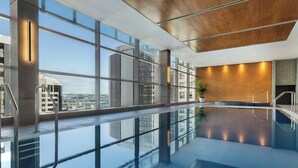 Une piscine intérieure moderne avec des murs en verre transparent, un plafond en bois et une vue sur les bâtiments de la ville.