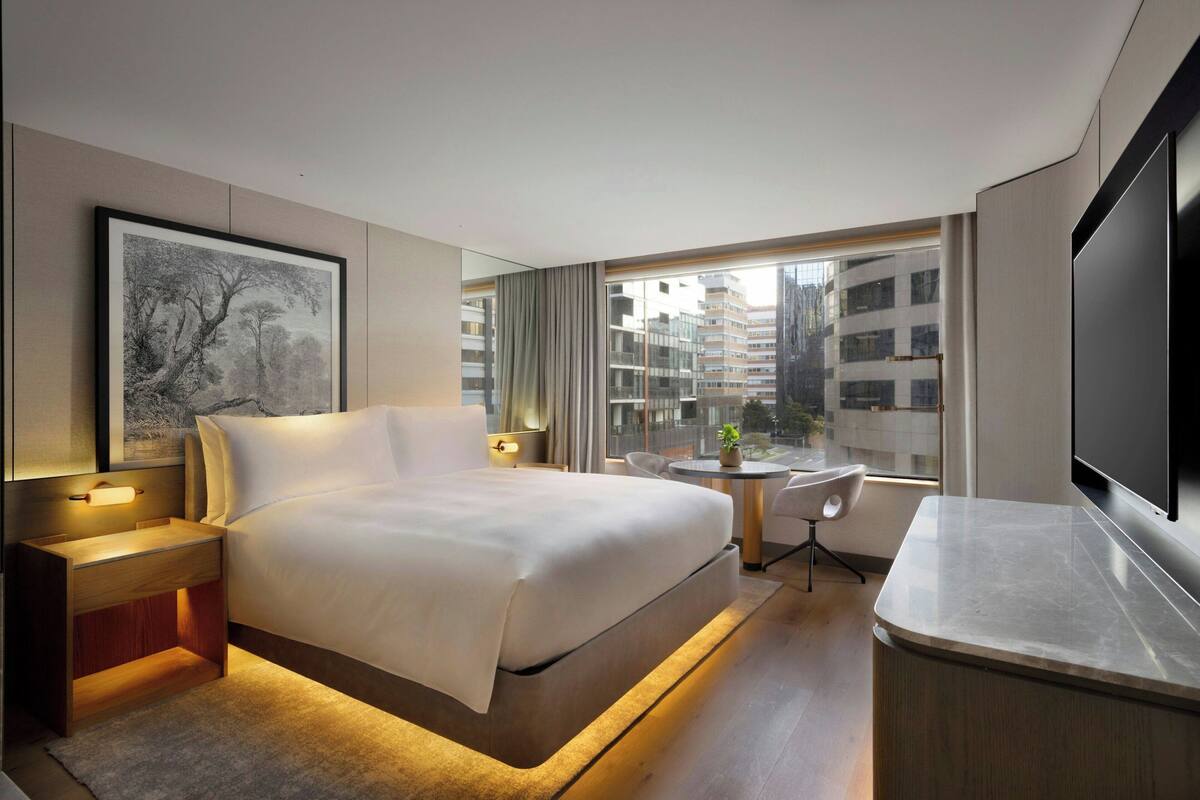 A modern hotel room with a large bed, bedside tables, a desk, a chair, a TV, and a window with a city view.