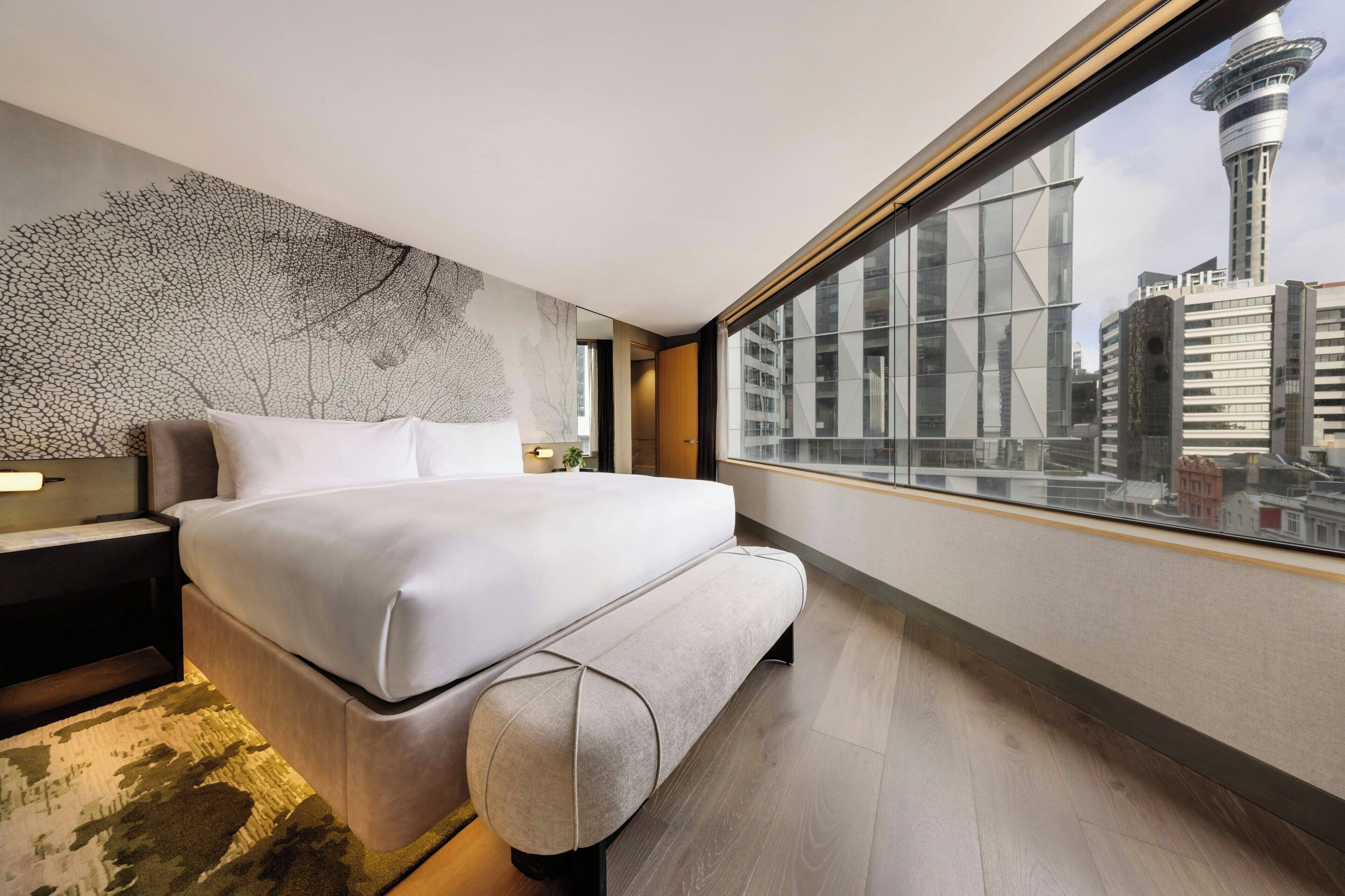 A modern hotel room with a large bed, a city view, and a unique wall art piece.