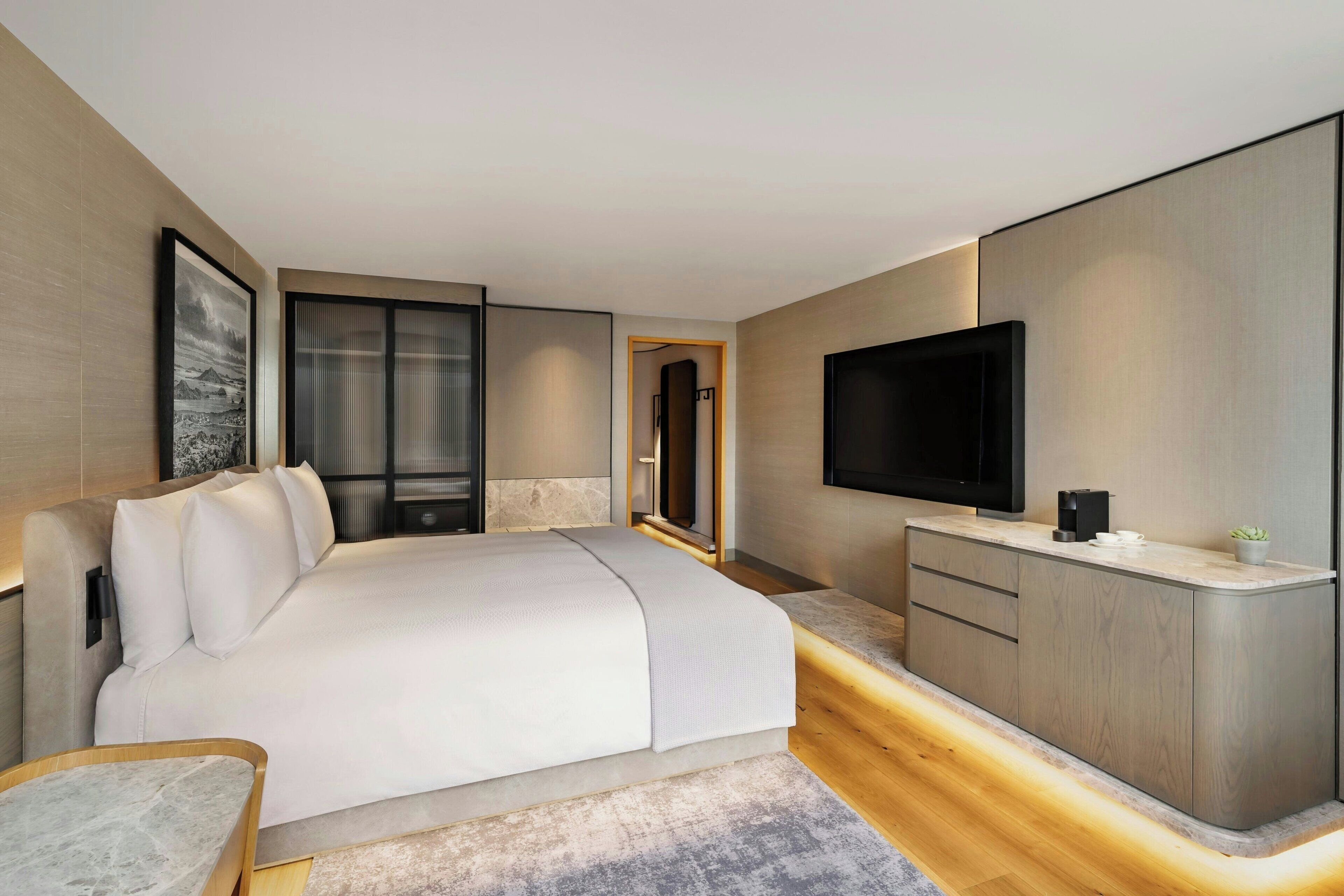 A modern hotel room with a large bed, a TV, a desk, and a view of the city.