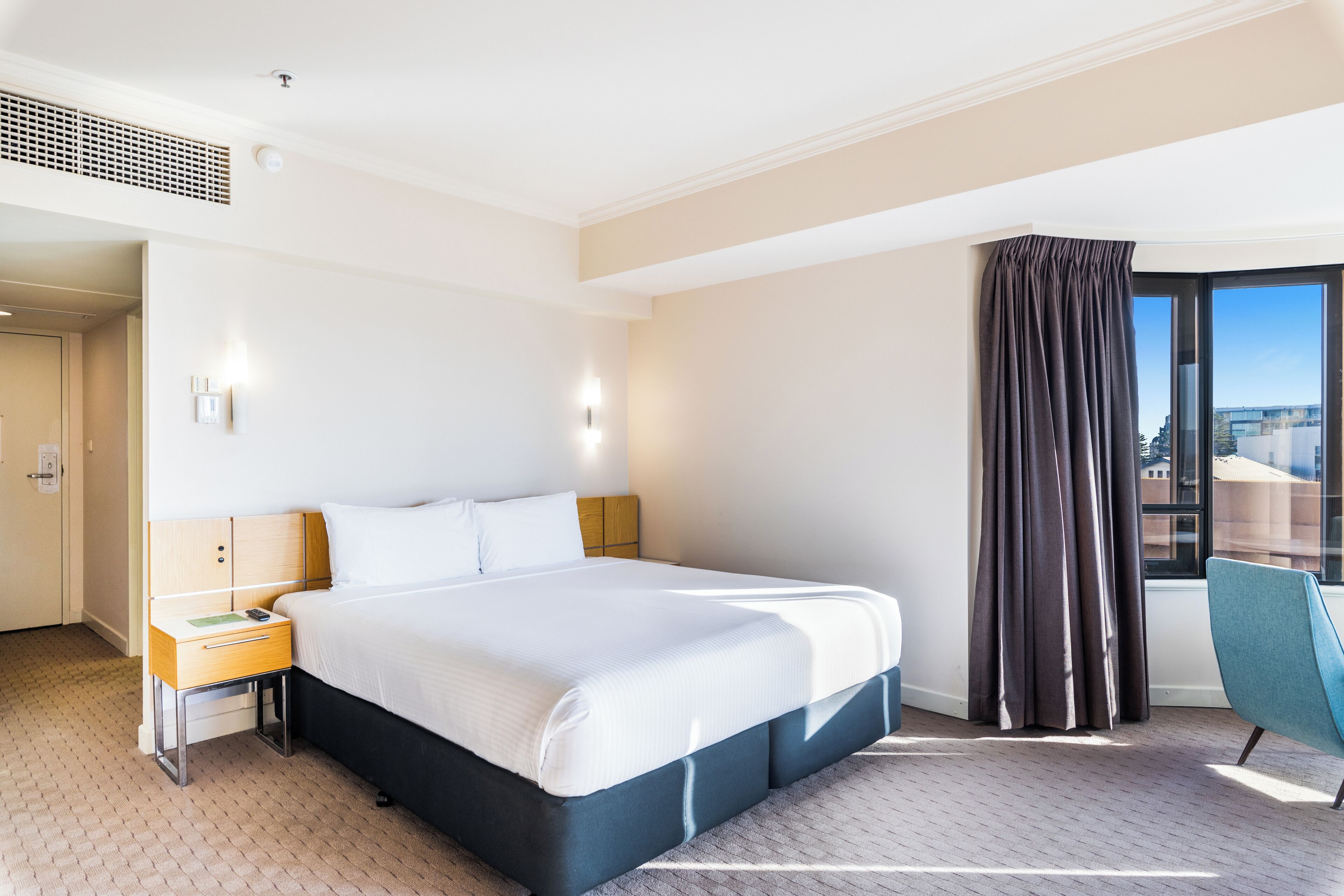 Habitación de hotel con una cama grande, una mesita de noche, una silla y vistas a la ciudad a través del ventanal.