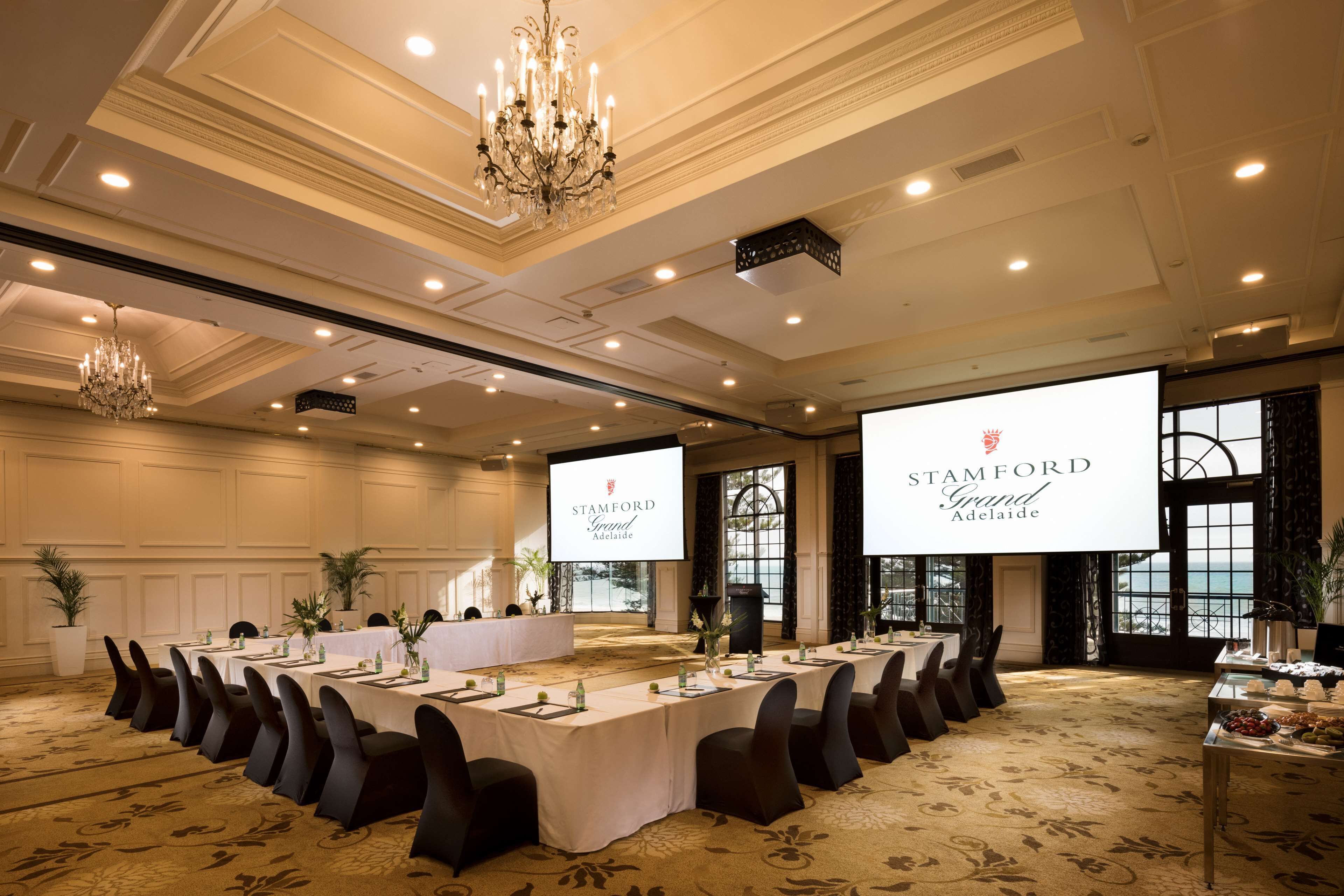 A conference room with a long table, chairs, a chandelier, and a screen displaying 'STAMFORD Grand Adelaide'.