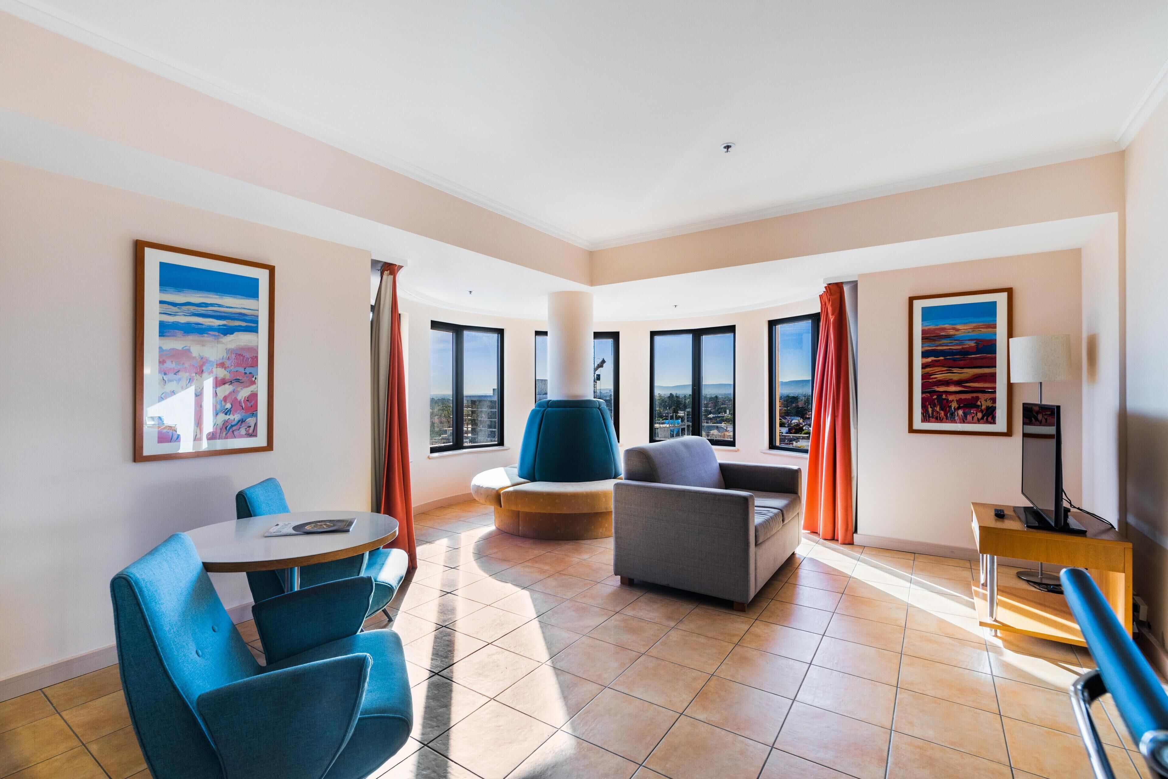 A hotel room with a large window, a round table, a sofa, and a TV.