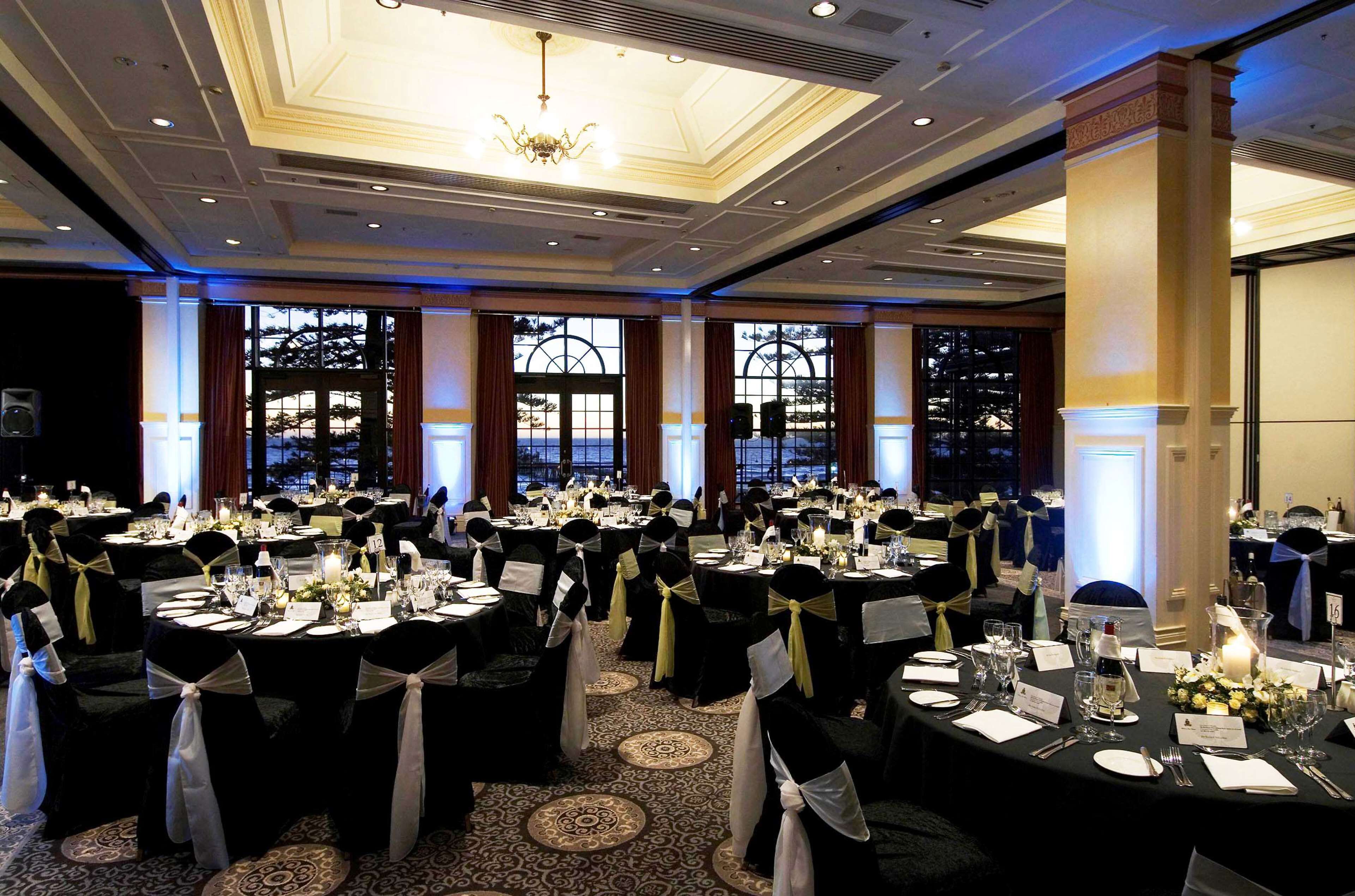 A banquet hall with round tables, black and white chairs, chandeliers, and large windows.