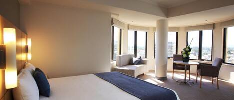Habitación de hotel con una cama grande, zona de estar con mesa y sillas, y ventanas amplias con vistas a la ciudad.