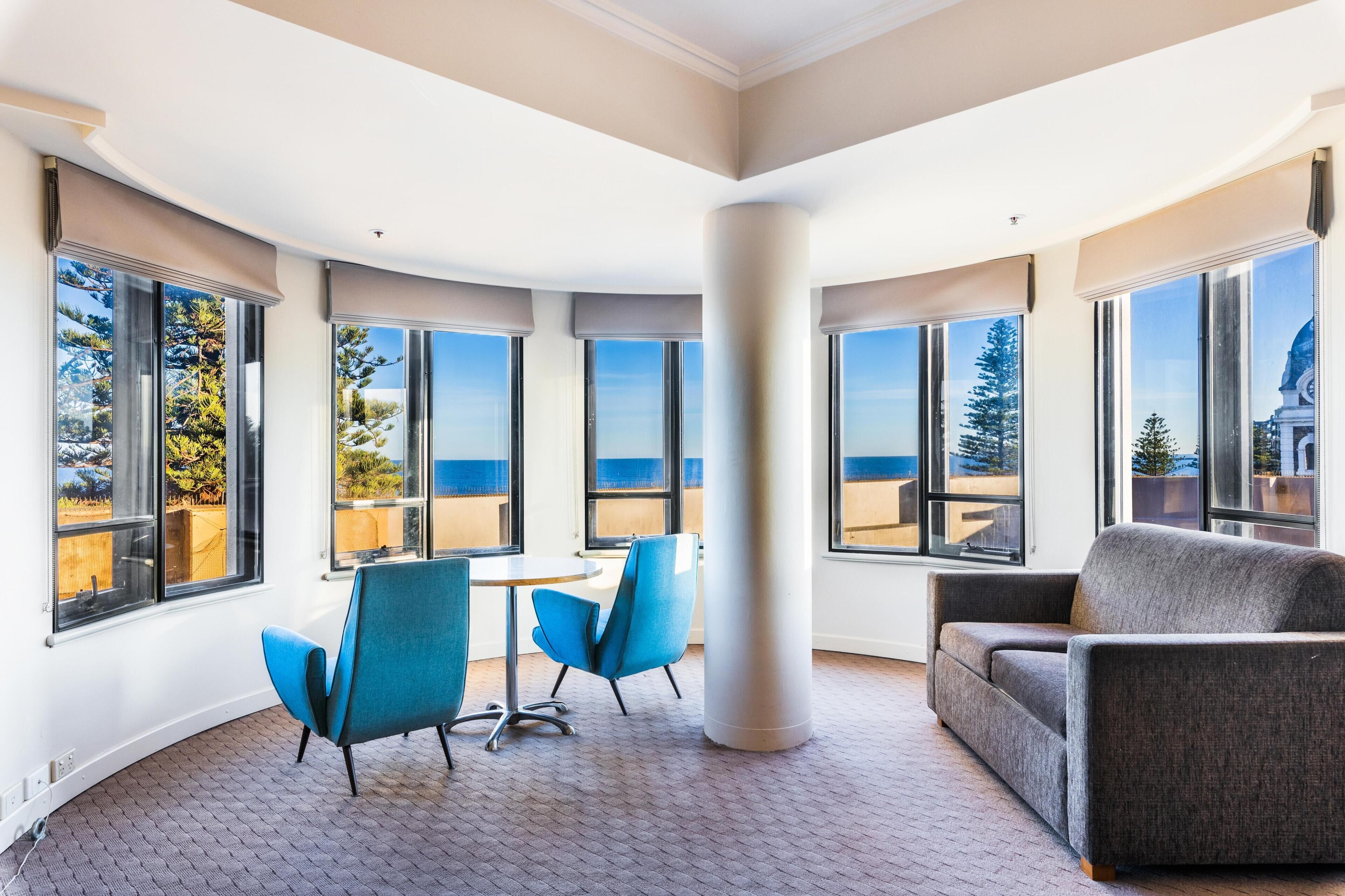 A hotel room with large windows, a round table, two blue chairs, a grey sofa, and a carpeted floor.