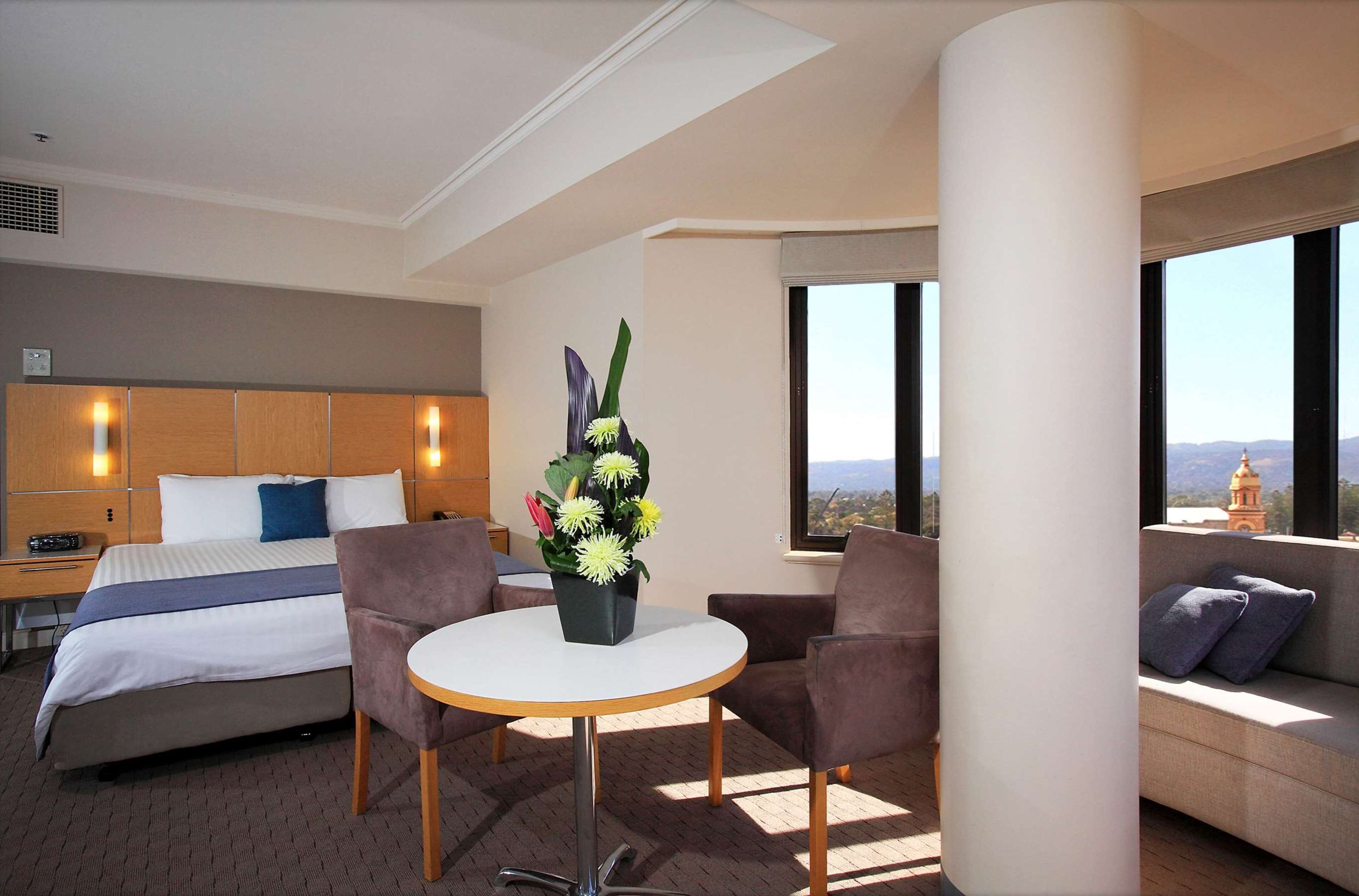 A hotel room with a bed, a round table with chairs, a sofa, and a large window with a view.