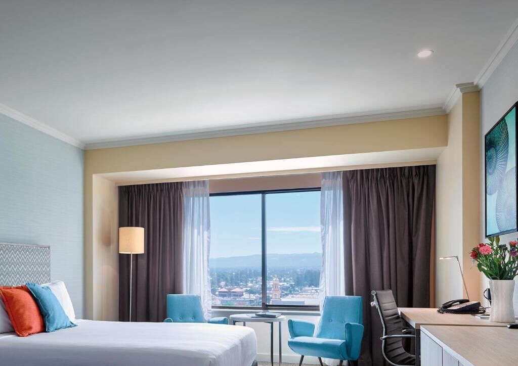 A hotel room with a bed, two blue chairs, a desk, and a large window with curtains.
