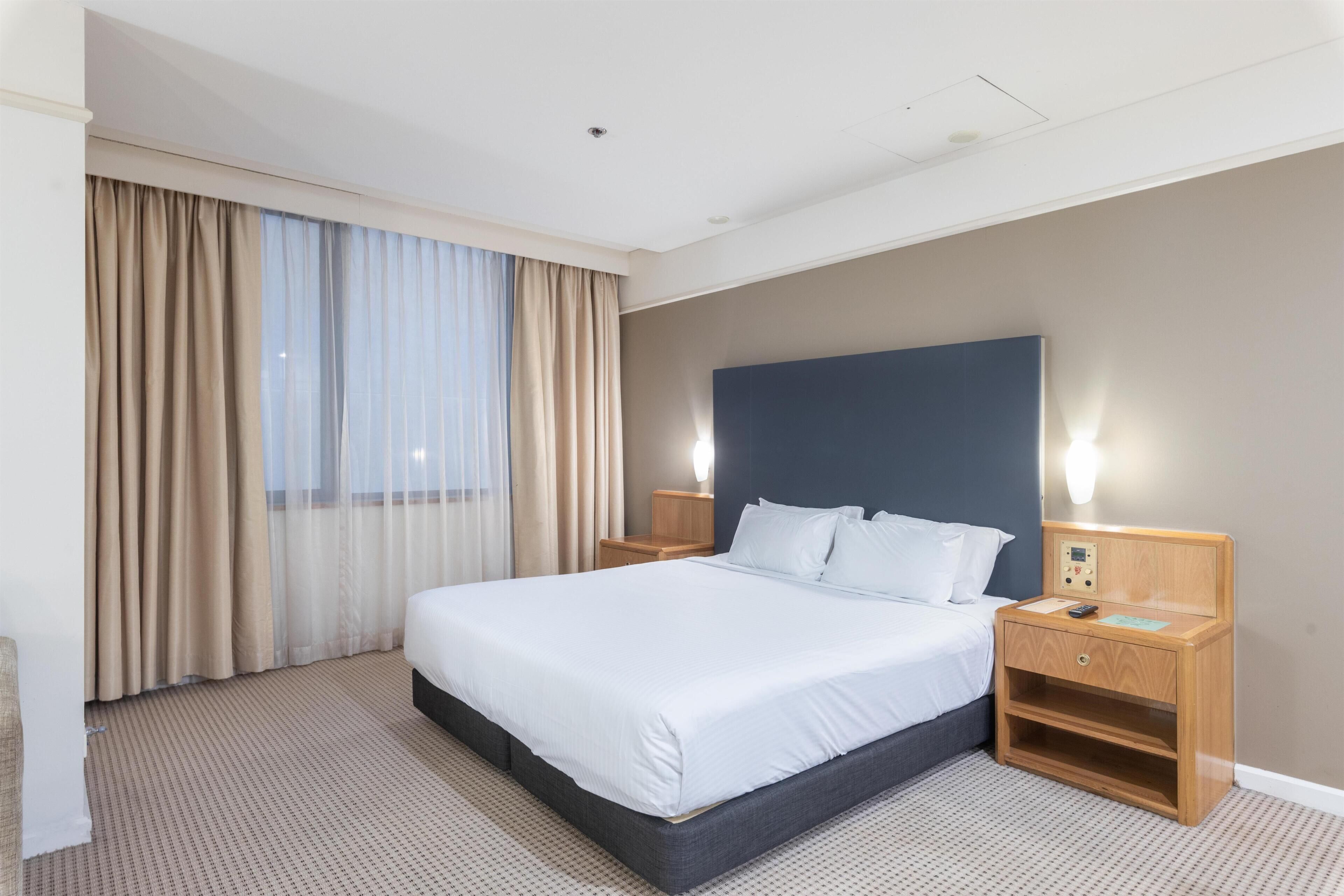 A hotel room with a large bed, a wooden nightstand, a wall-mounted control panel, and a window with curtains.