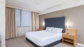 A hotel room with a large bed, a wooden nightstand, a wall-mounted control panel, and a window with curtains.