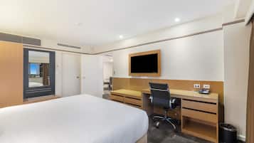 A hotel room with a bed, desk, chair, TV, and a trash bin.