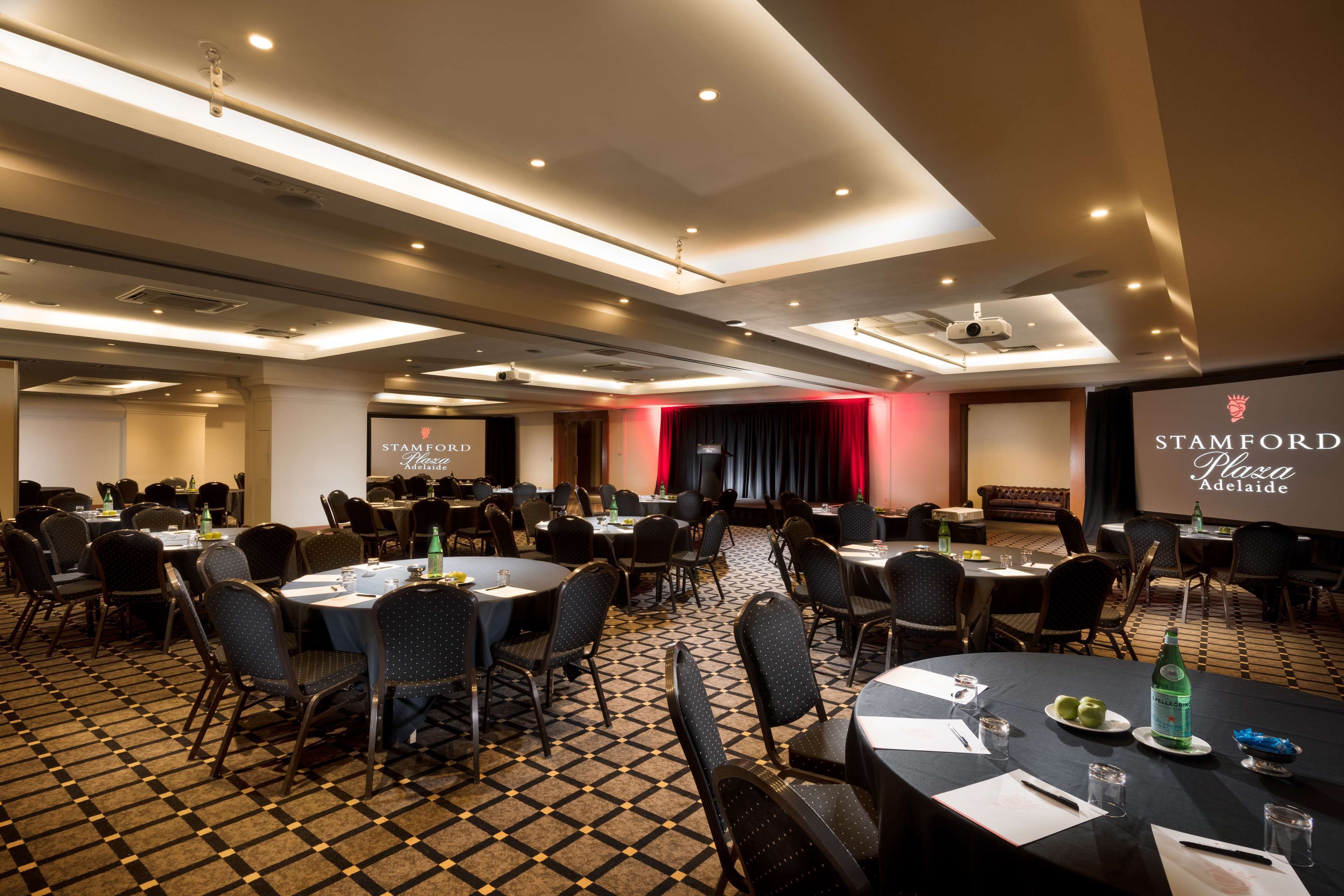 Un salón de conferencias con mesas, sillas y un escenario con una pantalla para proyector.