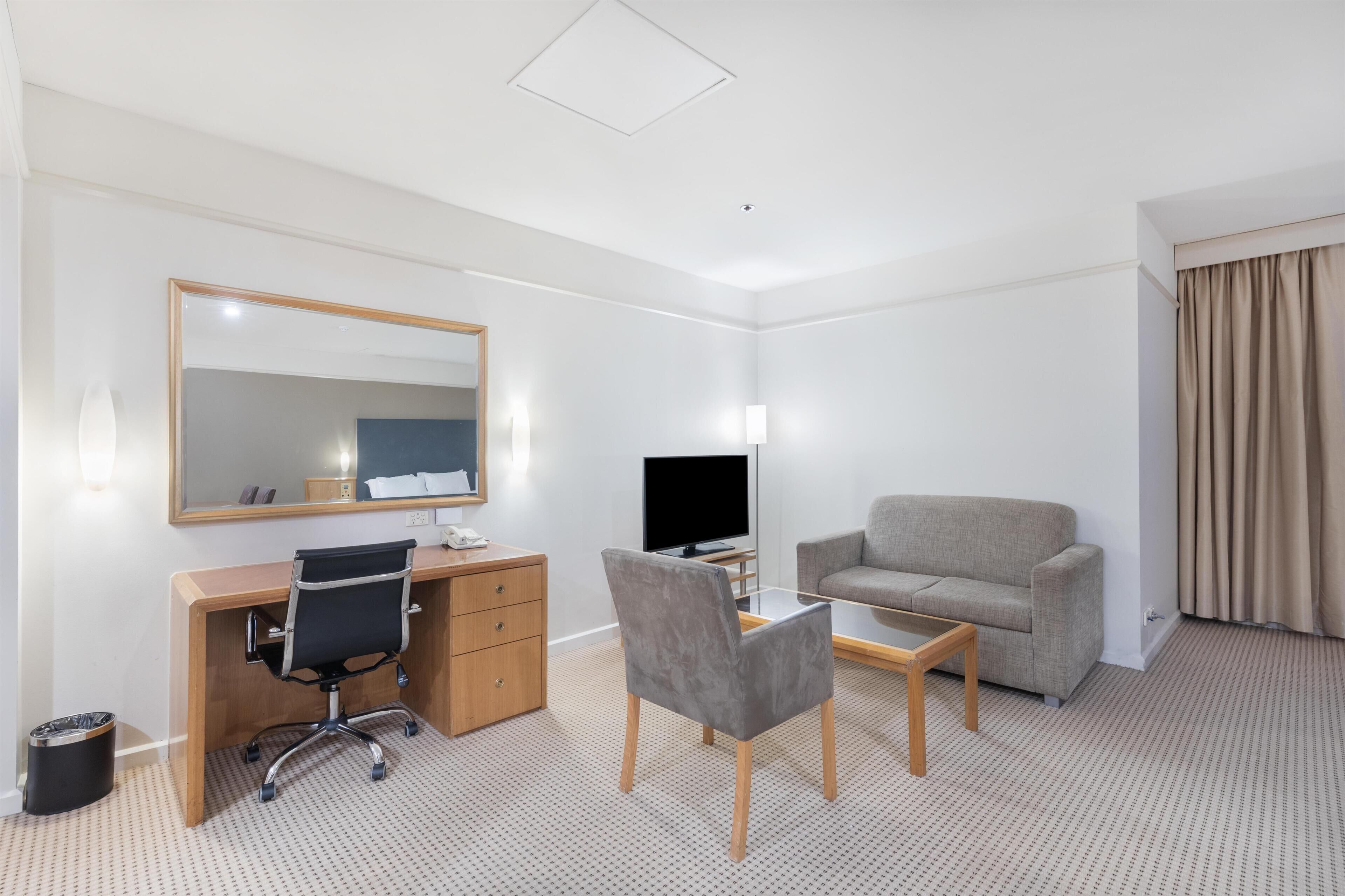A hotel room with a desk, chair, television, sofa, and a mirror.