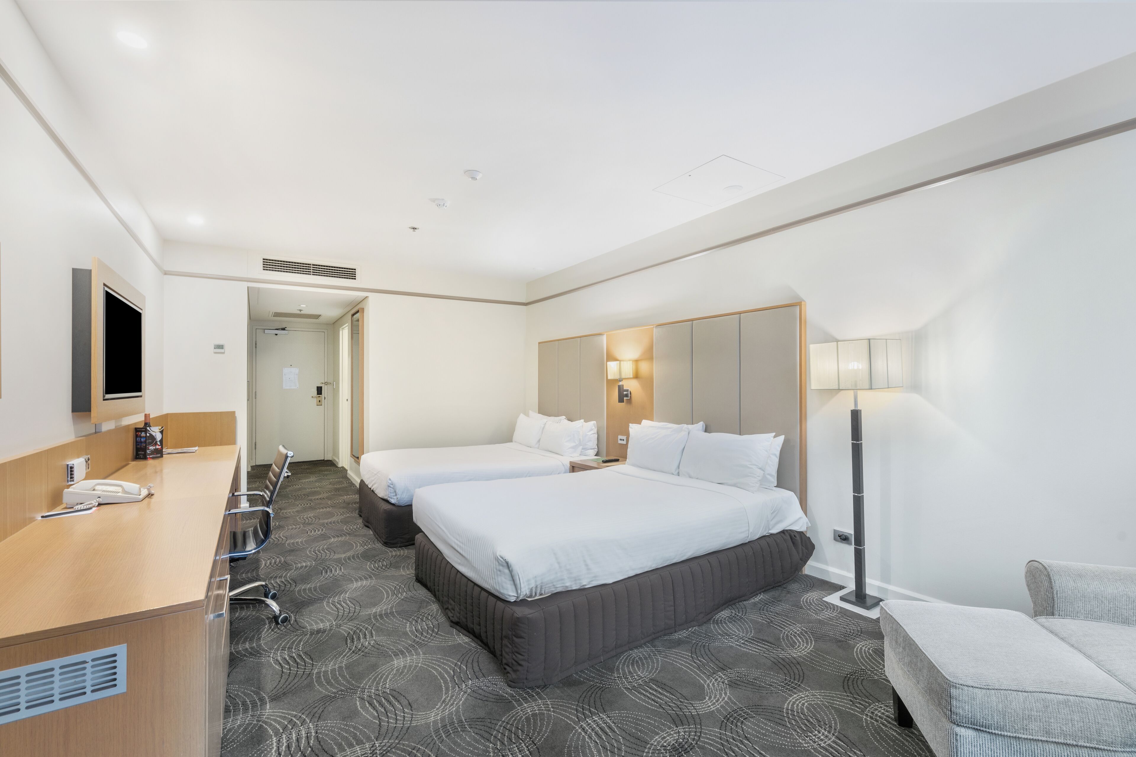 Une chambre d’hôtel avec deux lits, un bureau avec un téléphone, une chaise, un canapé, une lampe et un téléviseur à écran plat fixé au mur.