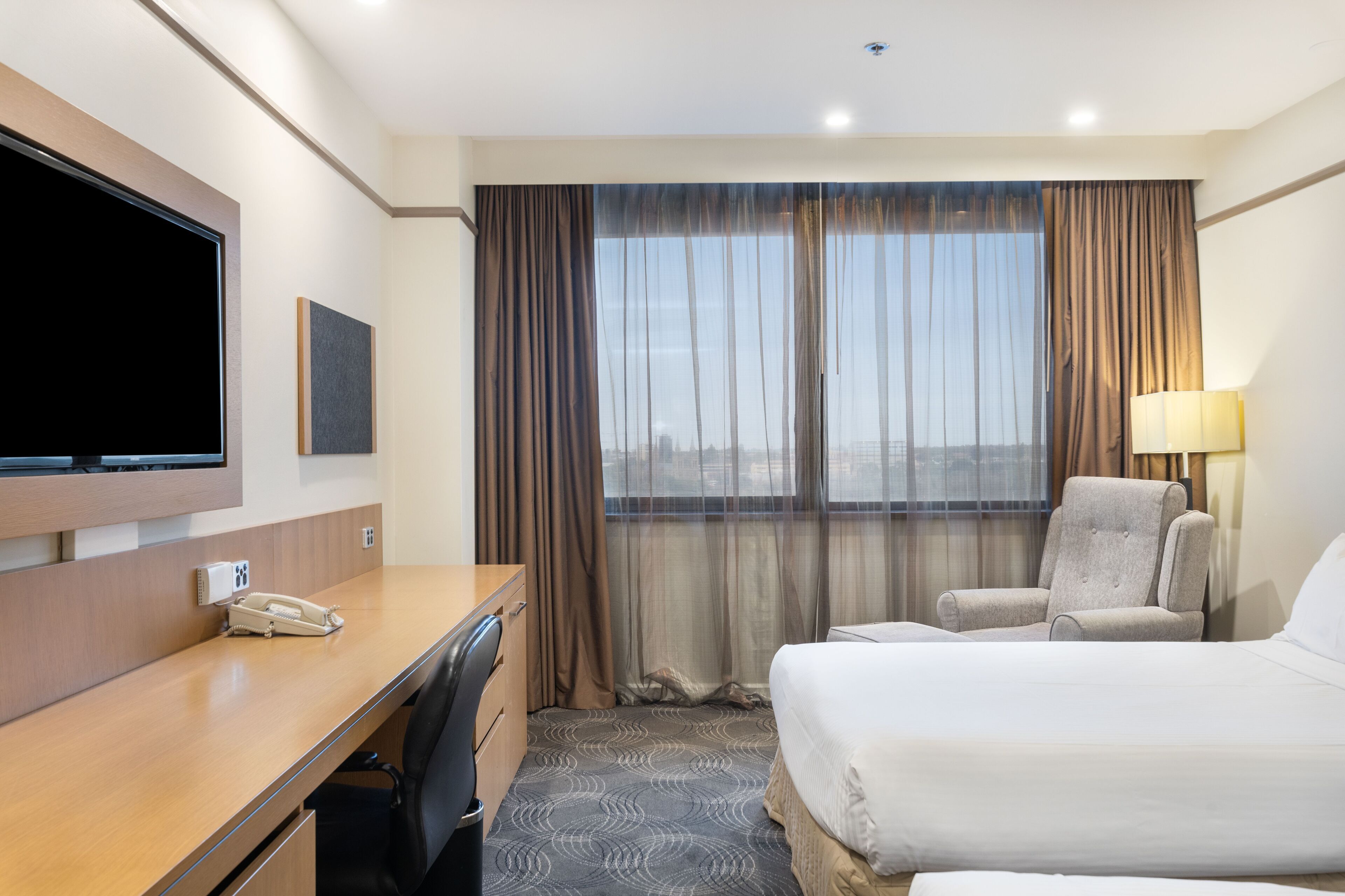 A hotel room with a bed, a desk with a telephone, a chair, a television mounted on the wall, and a window with curtains.