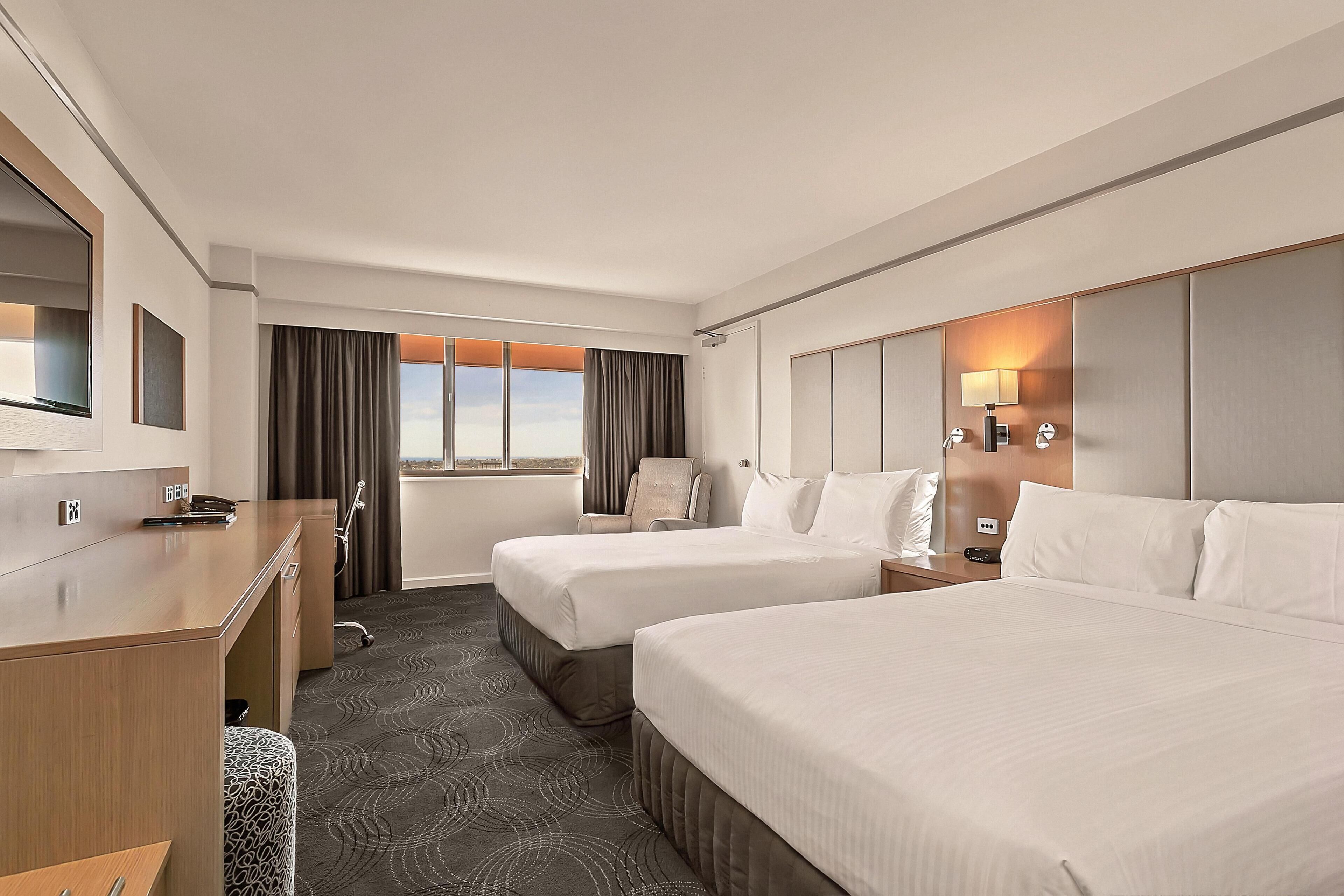 A hotel room with two beds, a desk, a chair, a bathroom, and a large window.